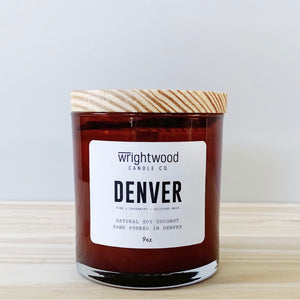 A candle in an amber colored glass jar with a wood lid is sitting on wood table with a neutral background. The candle has a white label with black text states the company name (Wrightwood Candle Co), scent name, scent notes, what it is made out of (soy-coconut wax), where it is hand poured (Denver) and item weight (9oz.)