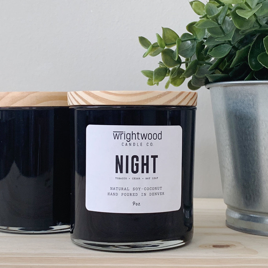 Two black jar candles are sitting on a wood table. The candle in the foreground has a white label and wood lid. The other candle has a wood lid, but no label visible. A metal planter with faux green foliage is on the right hand side. The background is a neutral color.