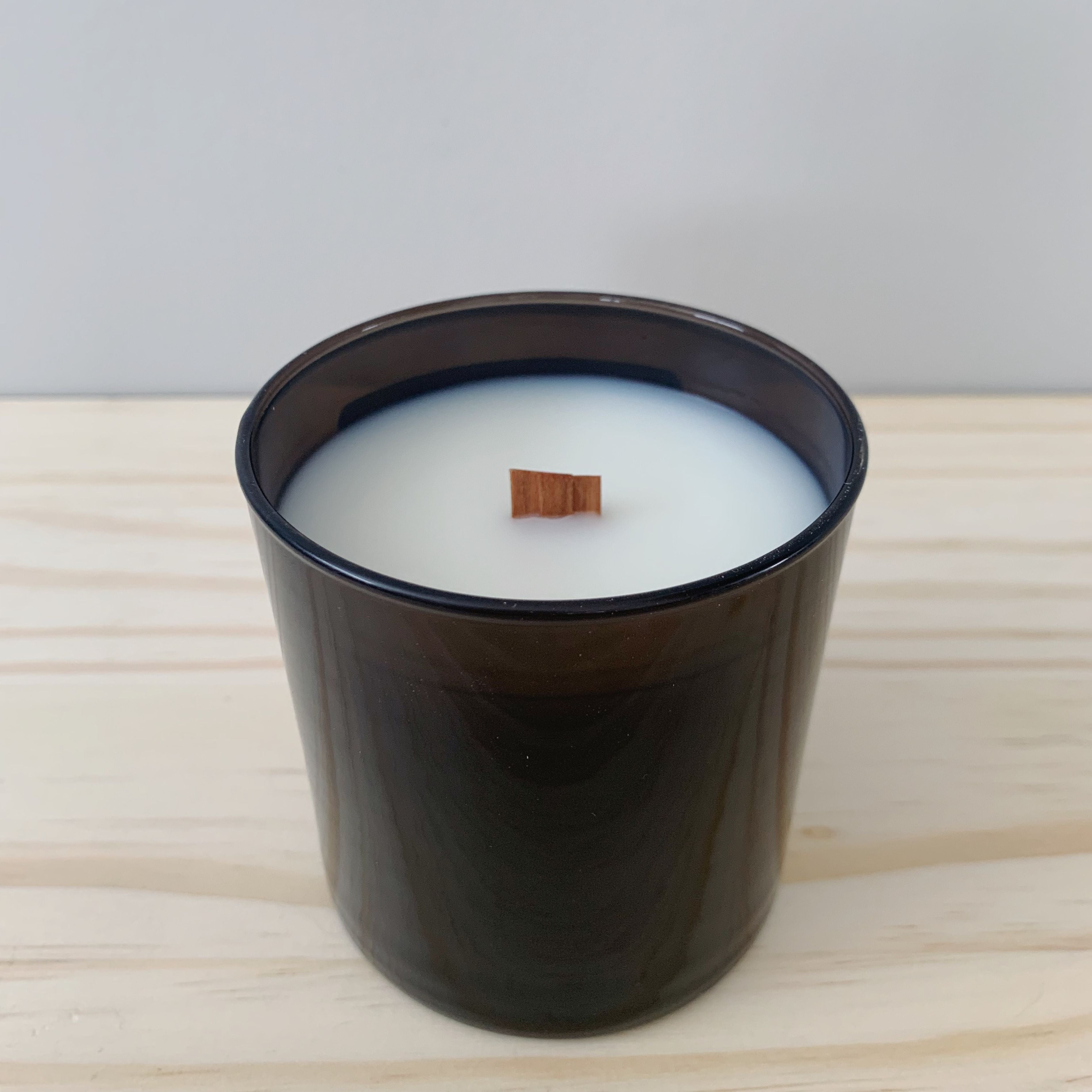 One black candle jar is sitting on top of a wood table with a neutral colored background. The candle has no lid or label. The wood wick is visible.