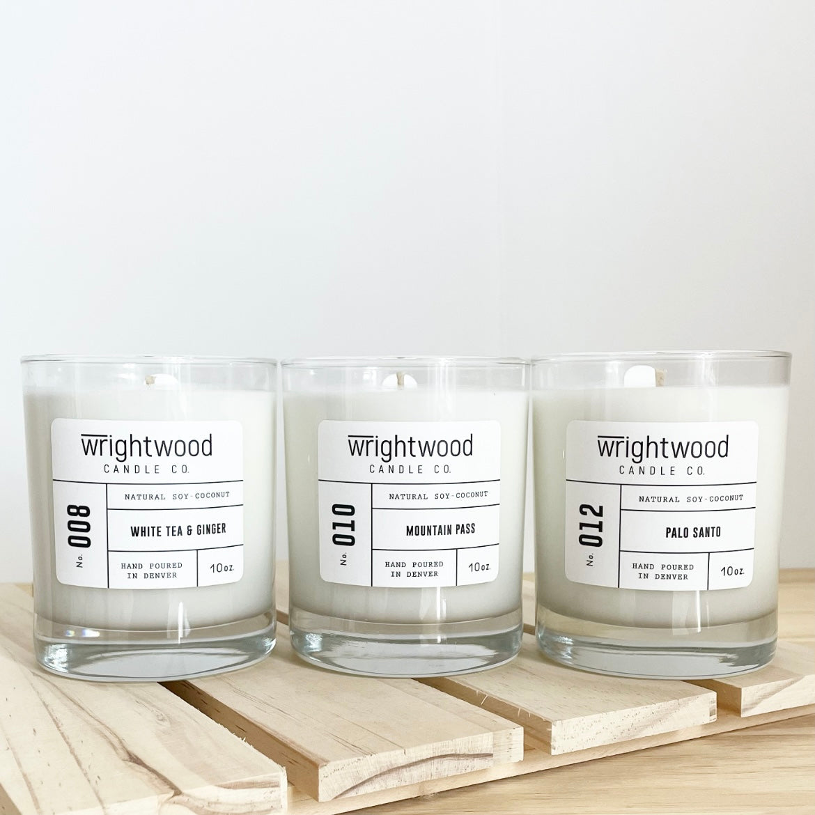 Three candles are sitting on top of a wood slatted board that is resting over a wood table. The background is neutral colored. The three candles are forward facing and have white labels with black text. The text includes the company name (Wrightwood Candle Co), scent name, what it is made out of (soy-coconut wax), where it is hand poured (Denver) and item weight (10oz.)