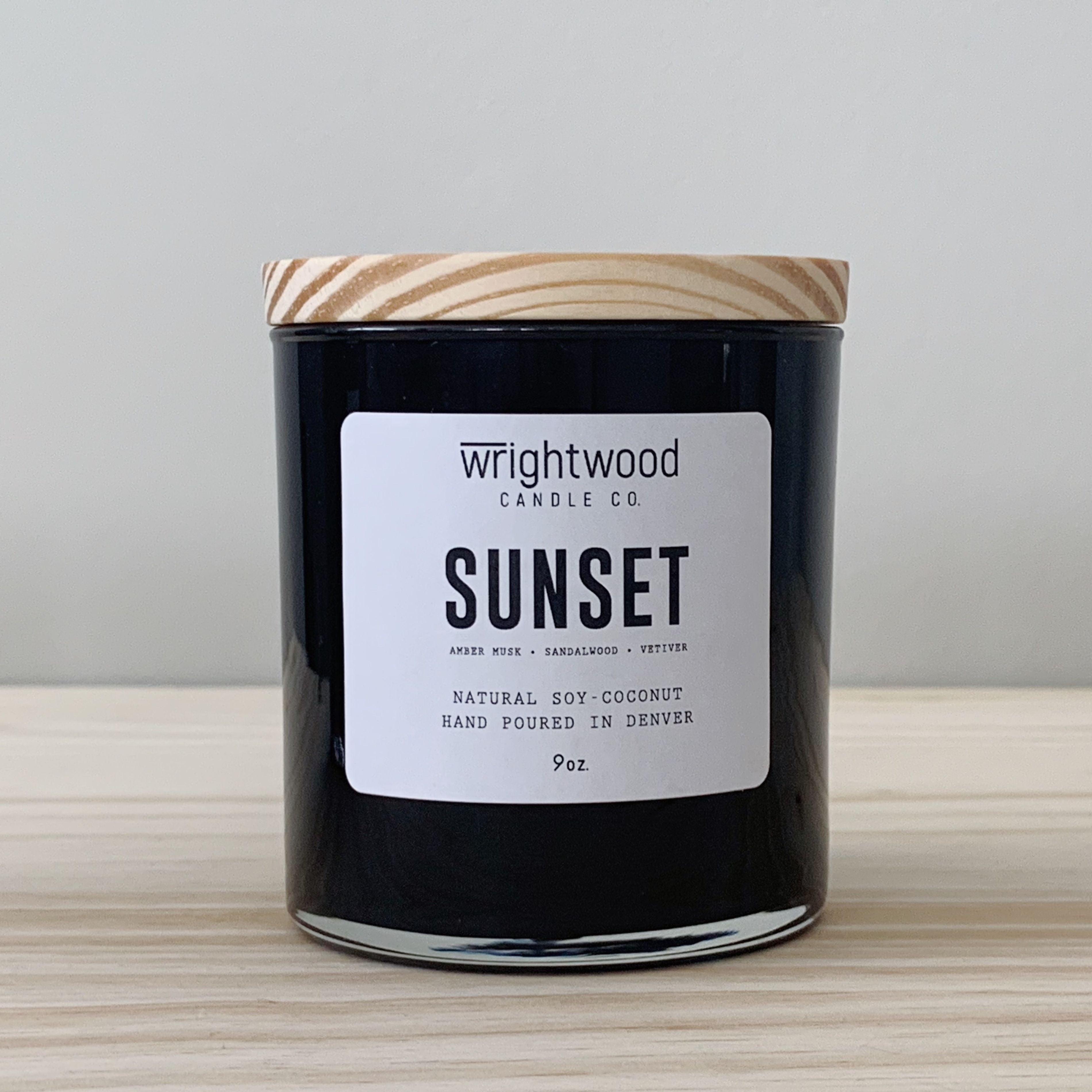 One black candle jar is sitting on top of a wood table with a neutral colored background. The candle has a wood lid and white label. The label has the scent name and company name visible. 