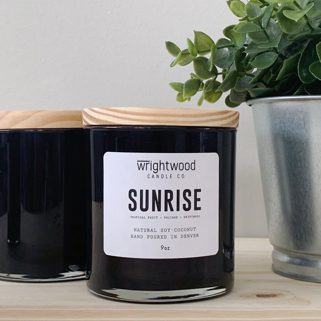 Two black jar candles are sitting on a wood table. The candle in the foreground has a white label and wood lid. The other candle has a wood lid, but no label visible. A metal planter with faux green foliage is on the right hand side. The background is a neutral color.