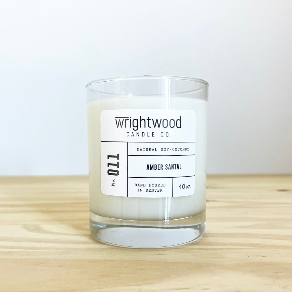 Container candle in a clear glass jar sitting on wood table with a white background. White label with black text states the company name (Wrightwood Candle Co), scent name, what it is made out of (soy-coconut wax), where it is hand poured (Denver) and item weight (10oz.)