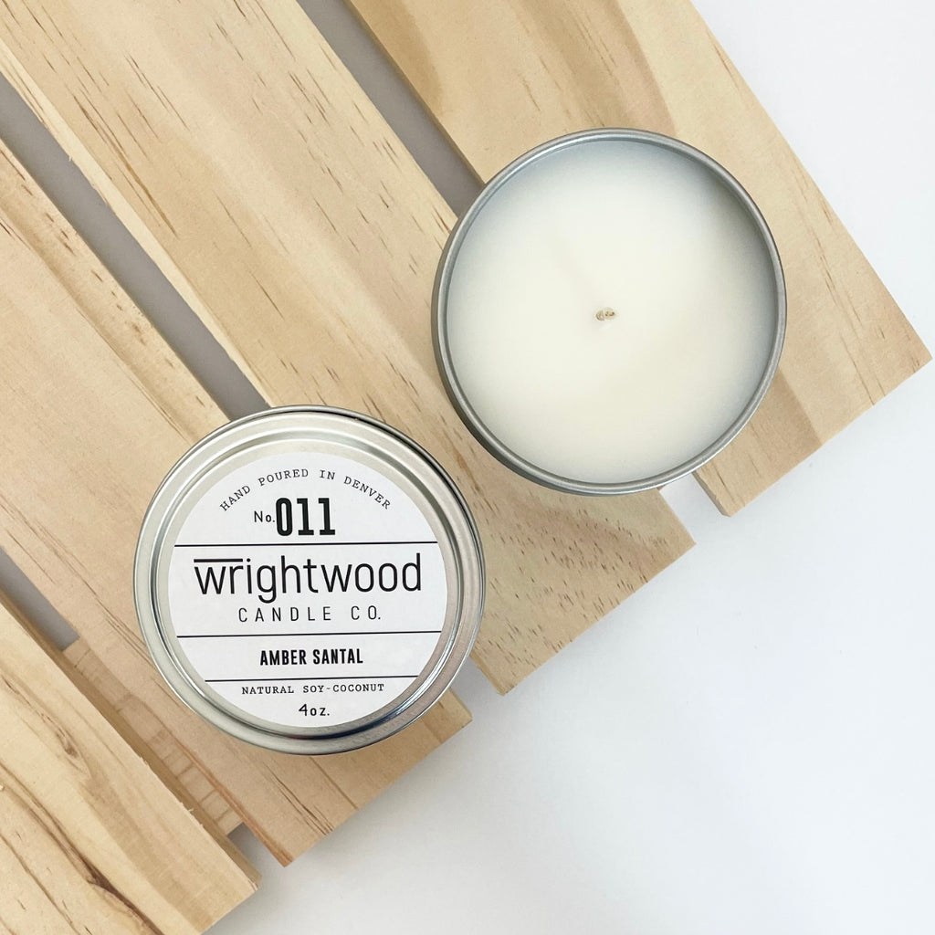 Two silver tins sit on top of a wood palleted board over a white table. The lower left tin on top of the board is showen with a white product label. The white product label includes the company name, scent name and size. The second candle on the wood board in the upper right is open showing the bare top of the candle.