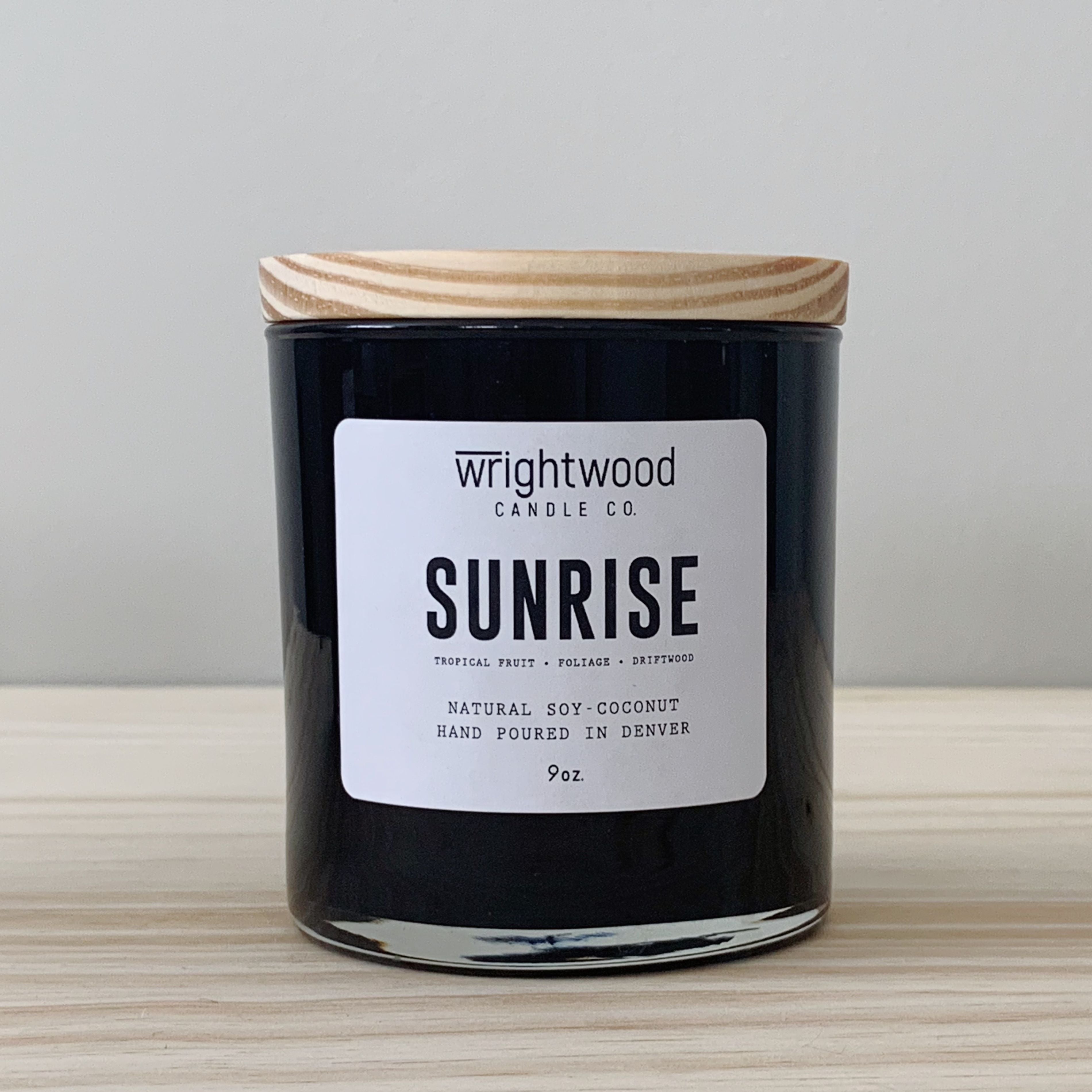 One black candle jar is sitting on top of a wood table with a neutral colored background. The candle has a wood lid and white label. The label has the scent name and company name visible. 