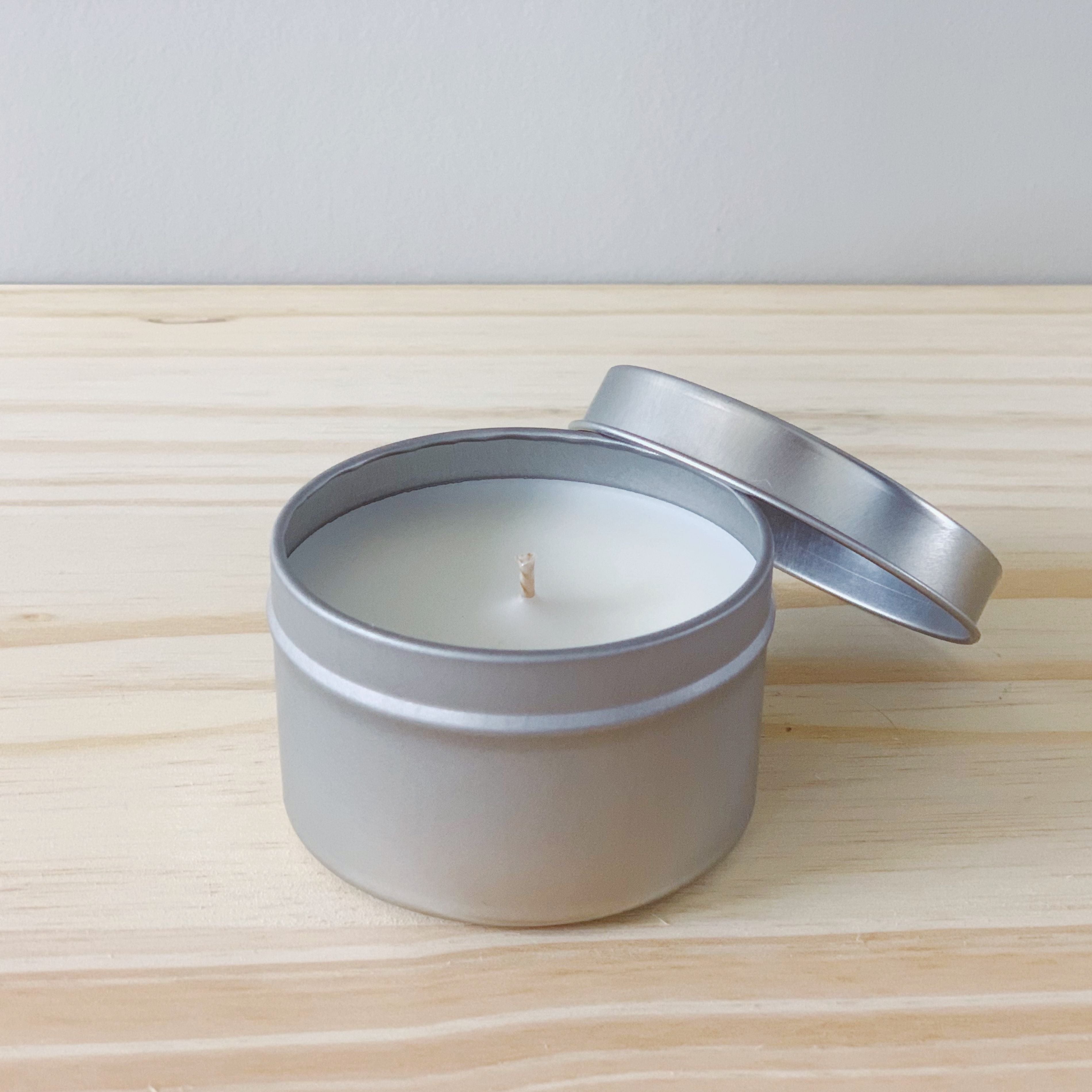 A single silver candle tin is shown on a wood table with a light colored background. The top of the candle is visible and the lid is resting on the back right of the tin.