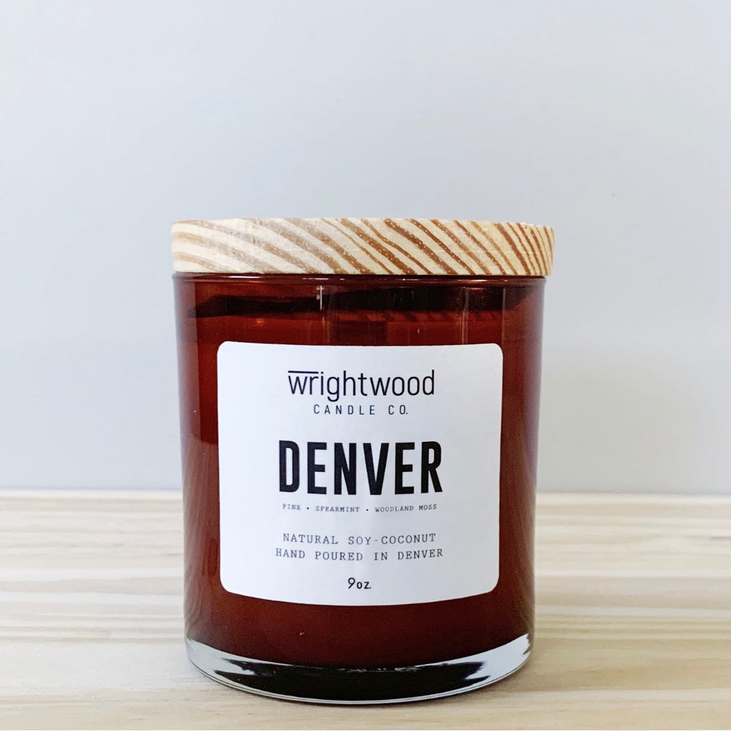 A candle in an amber colored glass jar with a wood lid is sitting on wood table with a neutral background. The candle has a white label with black text states the company name (Wrightwood Candle Co), scent name, scent notes, what it is made out of (soy-coconut wax), where it is hand poured (Denver) and item weight (9oz.)