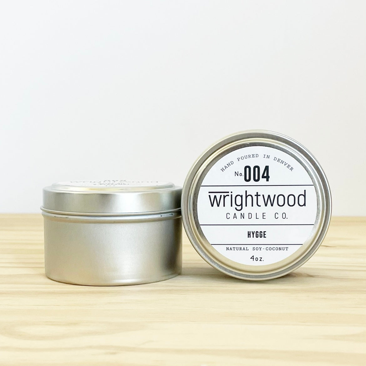 Two silver tins are on a wood table with a light colored background. The one on the left is upright while the one on the right has the lid facing outward with label visible. The label states the company name (Wrightwood Candle Co), scent name, what it is made out of (soy-coconut wax), where it is hand poured (Denver) and item weight (4oz.)
