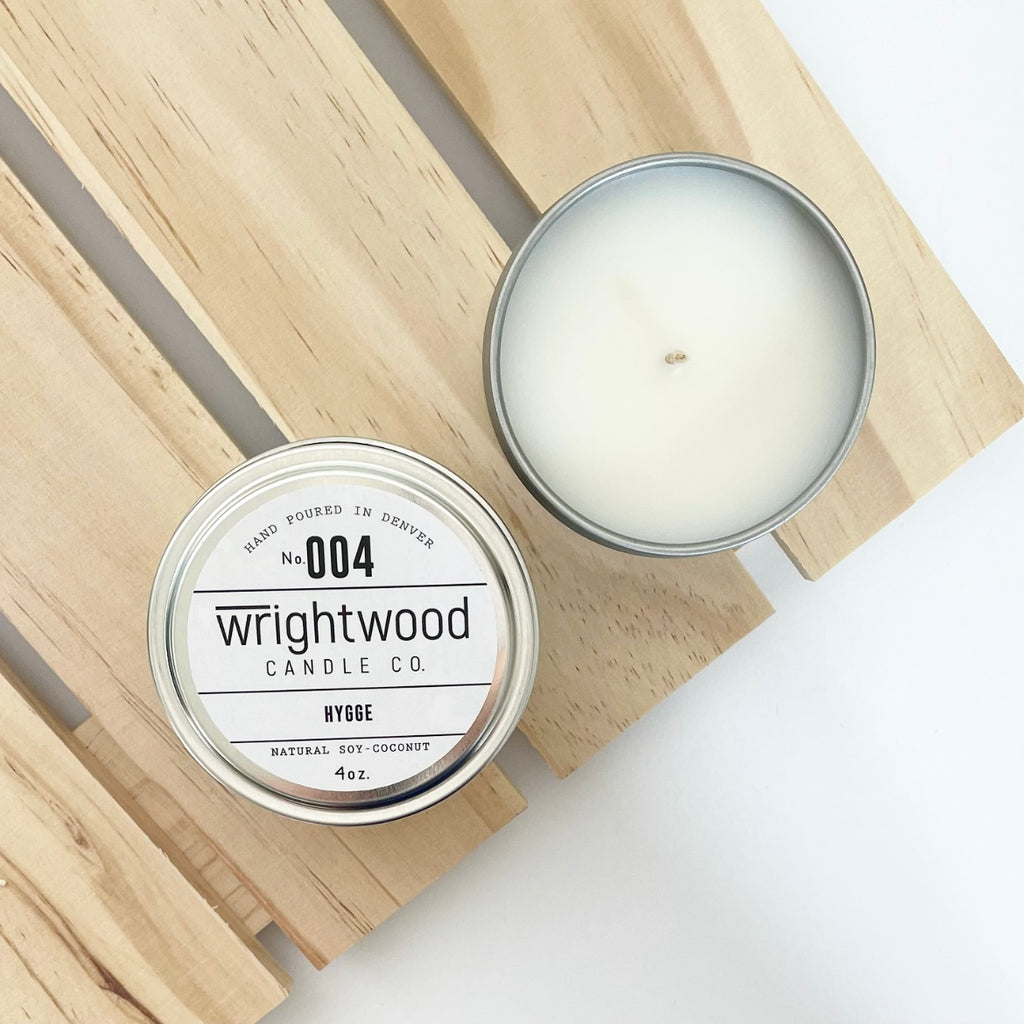 Two silver tins sit on top of a wood palleted board over a white table. The lower left tin on top of the board is showen with a white product label. The white product label includes the company name, scent name and size. The second candle on the wood board in the upper right is open showing the bare top of the candle.