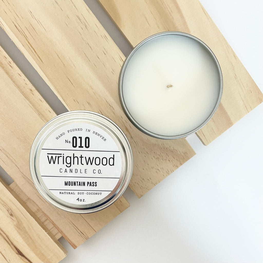 Two silver tins sit on top of a wood palleted board over a white table. The lower left tin on top of the board is showen with a white product label. The white product label includes the company name, scent name and size. The second candle on the wood board in the upper right is open showing the bare top of the candle.