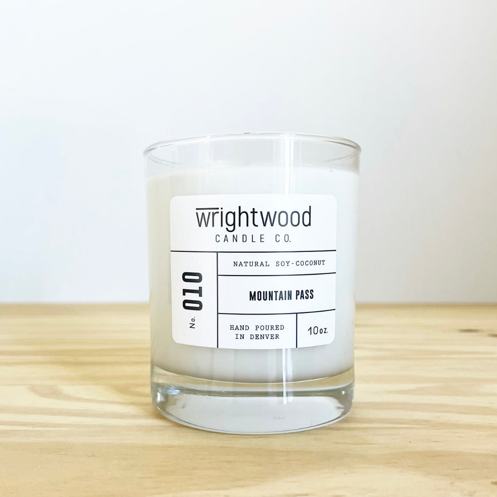 Container candle in a clear glass jar sitting on wood table with a white background. White label with black text states the company name (Wrightwood Candle Co), scent name, what it is made out of (soy-coconut wax), where it is hand poured (Denver) and item weight (10oz.)