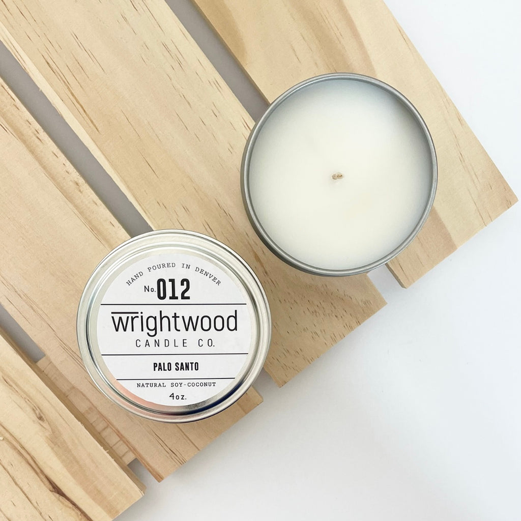 Two silver tins sit on top of a wood palleted board over a white table. The lower left tin on top of the board is showen with a white product label. The white product label includes the company name, scent name and size. The second candle on the wood board in the upper right is open showing the bare top of the candle.