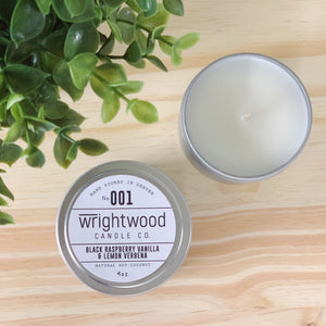 Two silver tins are sitting on top of a wood table. Faux green foliage is seen in the top left corner. The top right candle tin is open. Top of the candle and candle wick is visible. The bottom left candle tin has a white label with black text. The label states the company name (Wrightwood Candle Co), scent name, what it is made out of (soy-coconut wax), where it is hand poured (Denver) and item weight (4oz.)