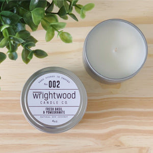Two silver tins are sitting on top of a wood table. Faux green foliage is seen in the top left corner. The top right candle tin is open. Top of the candle and candle wick is visible. The bottom left candle tin has a white label with black text. The label states the company name (Wrightwood Candle Co), scent name, what it is made out of (soy-coconut wax), where it is hand poured (Denver) and item weight (4oz.)