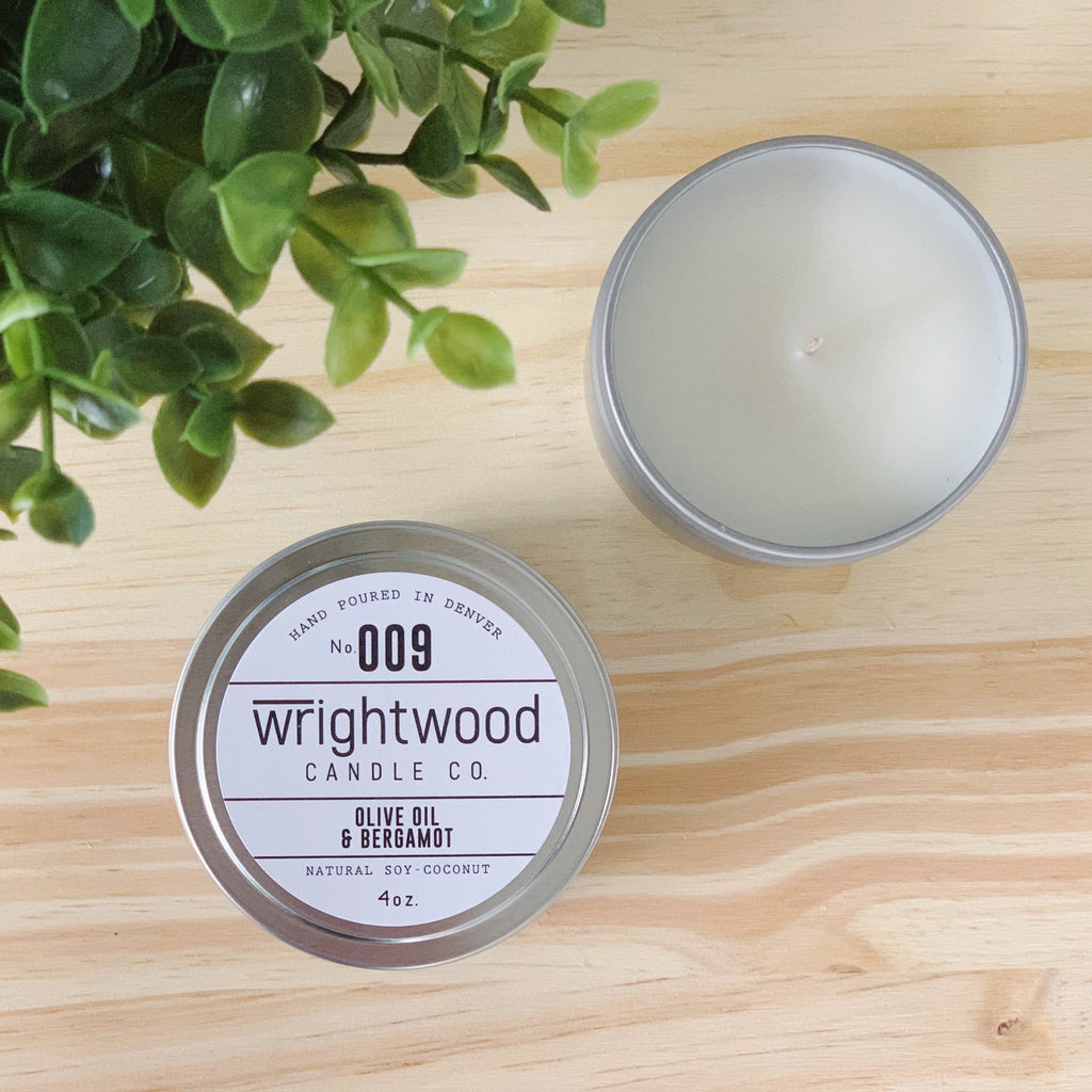 Two silver tins are sitting on top of a wood table. Faux green foliage is seen in the top left corner. The top right candle tin is open. Top of the candle and candle wick is visible. The bottom left candle tin has a white label with black text. The label states the company name (Wrightwood Candle Co), scent name, what it is made out of (soy-coconut wax), where it is hand poured (Denver) and item weight (4oz.)